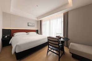 Une chambre d’hôtel comprenant un lit, un bureau, une chaise, une table de chevet, une lampe et une fenêtre avec des rideaux.