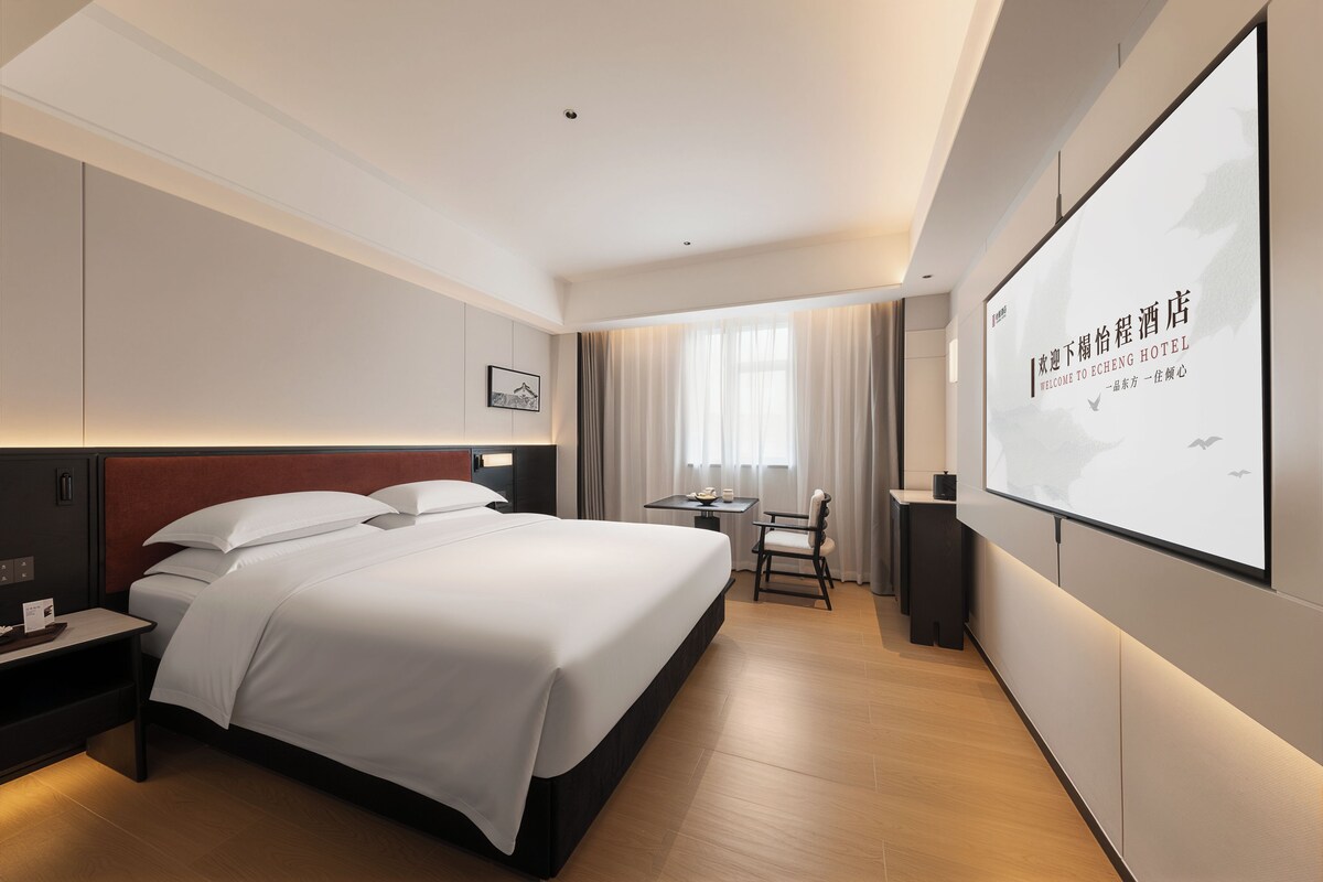 Une chambre d’hôtel moderne avec un grand lit, un bureau et une chaise. Un grand écran affiche du texte.