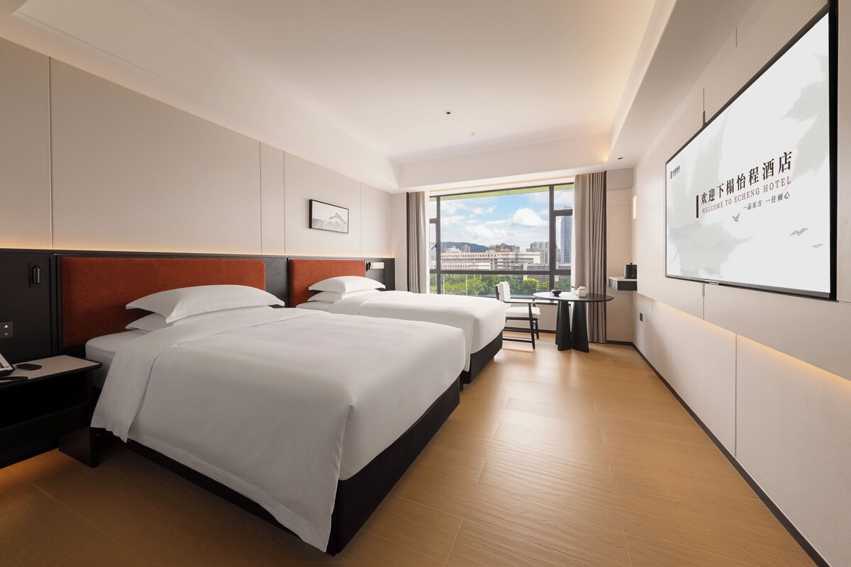 Une chambre d’hôtel avec deux lits, un bureau et une grande fenêtre donnant sur la ville.