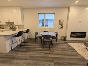 A modern kitchen with white cabinets, a dining area with a round table and chairs, a window with a view, and a fireplace.