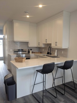 A modern kitchen with white cabinets, a stainless steel range, a large island with bar stools, and a black trash bin.