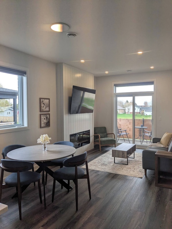 A modern living room with a fireplace, a dining area with a round table and chairs, and a view of the outdoors through large windows.