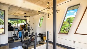 Et fitnessrum med træningsudstyr, en ventilator i loftet og store vinduer med udsigt til grønne områder.