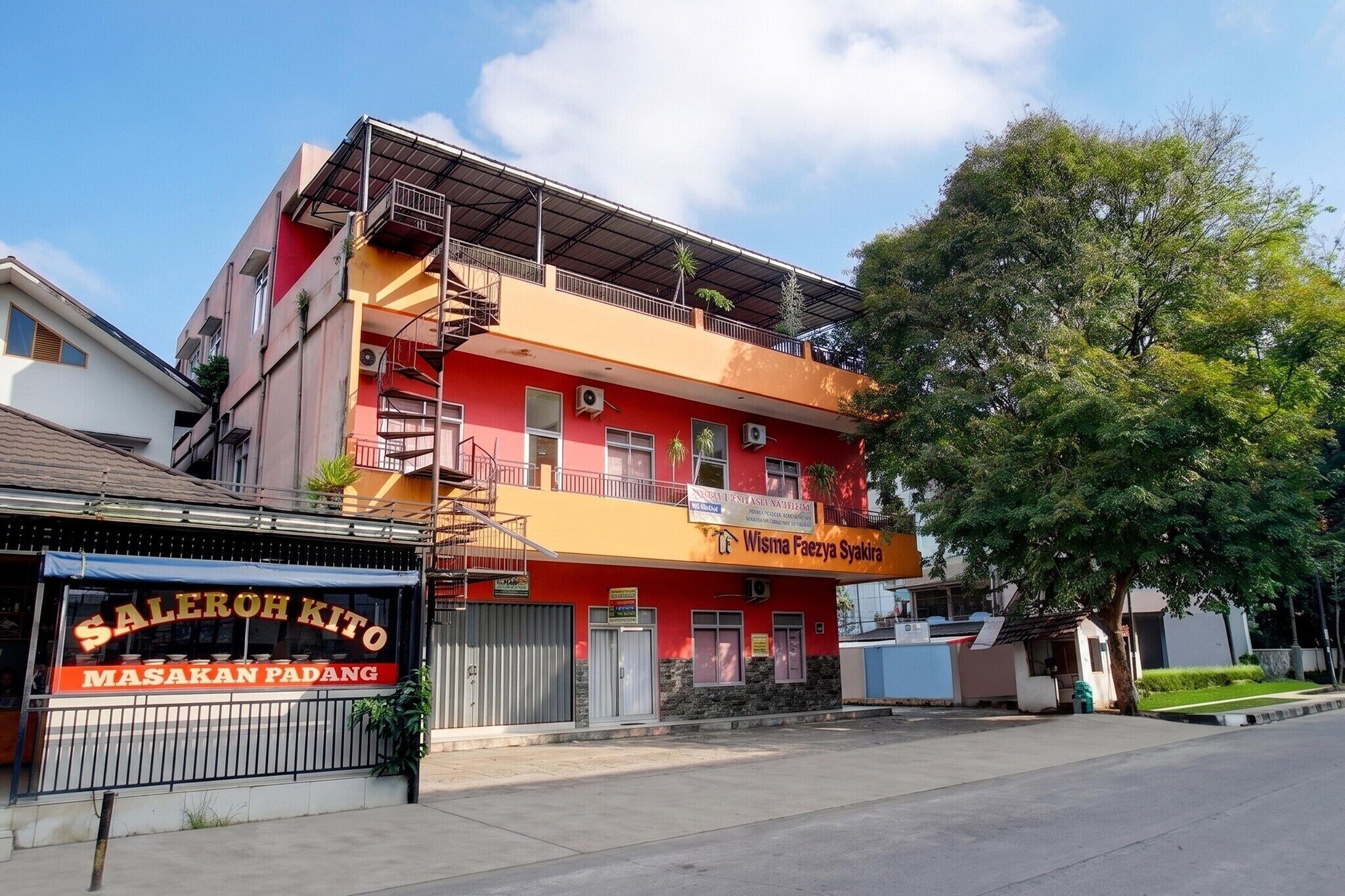 A multi-story building with a red and yellow facade, a visible balcony, and a sign for 'Wisma Faezya Syakira.'