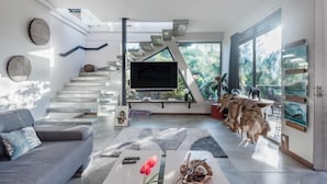 A modern living room with a flat-screen TV, a sofa, a coffee table with decorative items, a staircase, and a large window with a view of greenery.