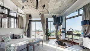 A modern hotel room with a large bed, a desk, a chair, a sofa, a large window, and a view of the city.