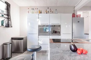 A modern kitchen with stainless steel appliances, a trash bin, and a marble countertop with vegetables.