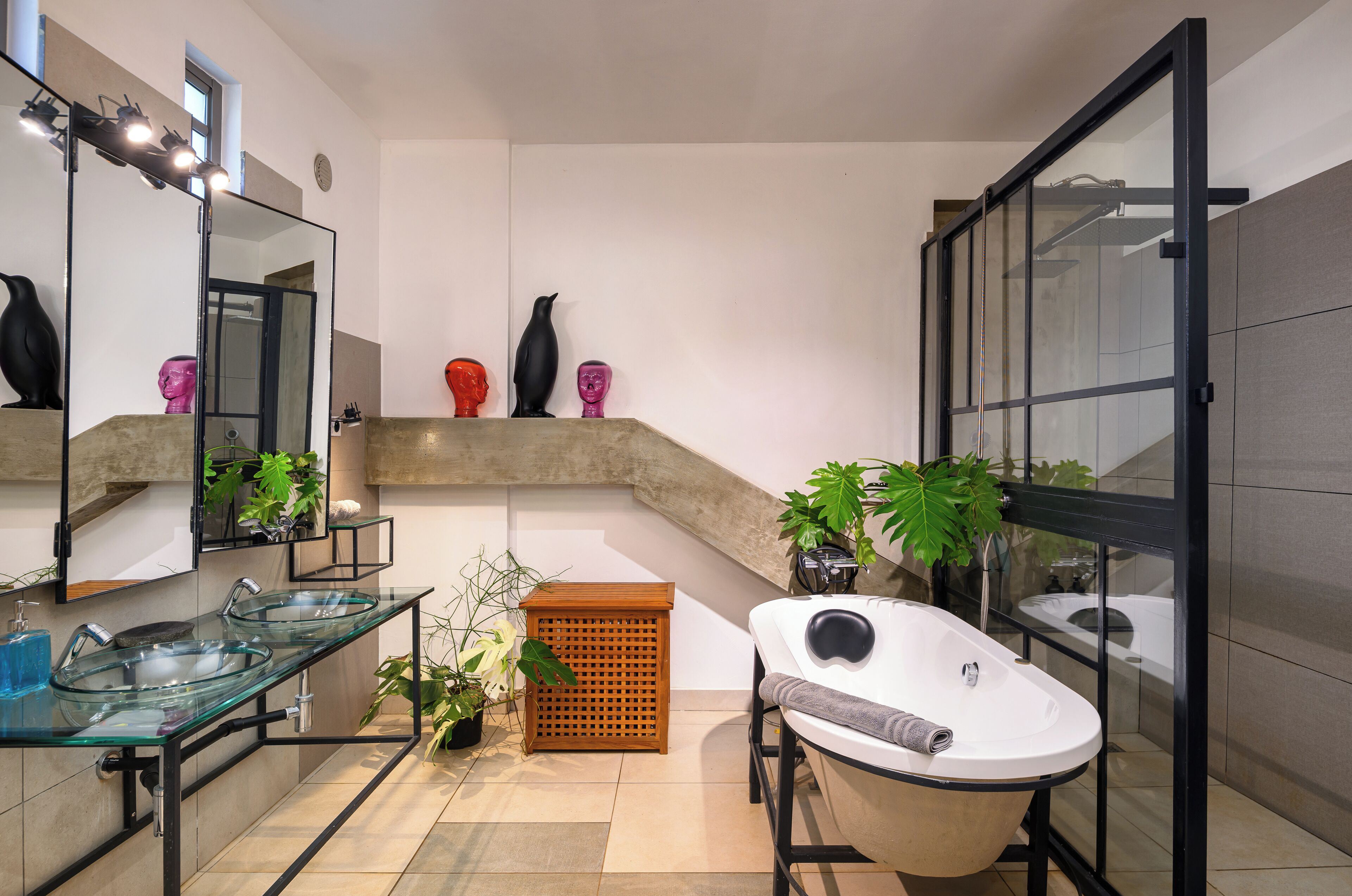 Un baño moderno con un lavabo de vidrio, una bañera independiente, un espejo con iluminación y un pequeño estante de madera con objetos decorativos.