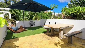 Eine Terrasse mit Tisch, Bank, Kunstgrras und einem großen Sonnenschirm.