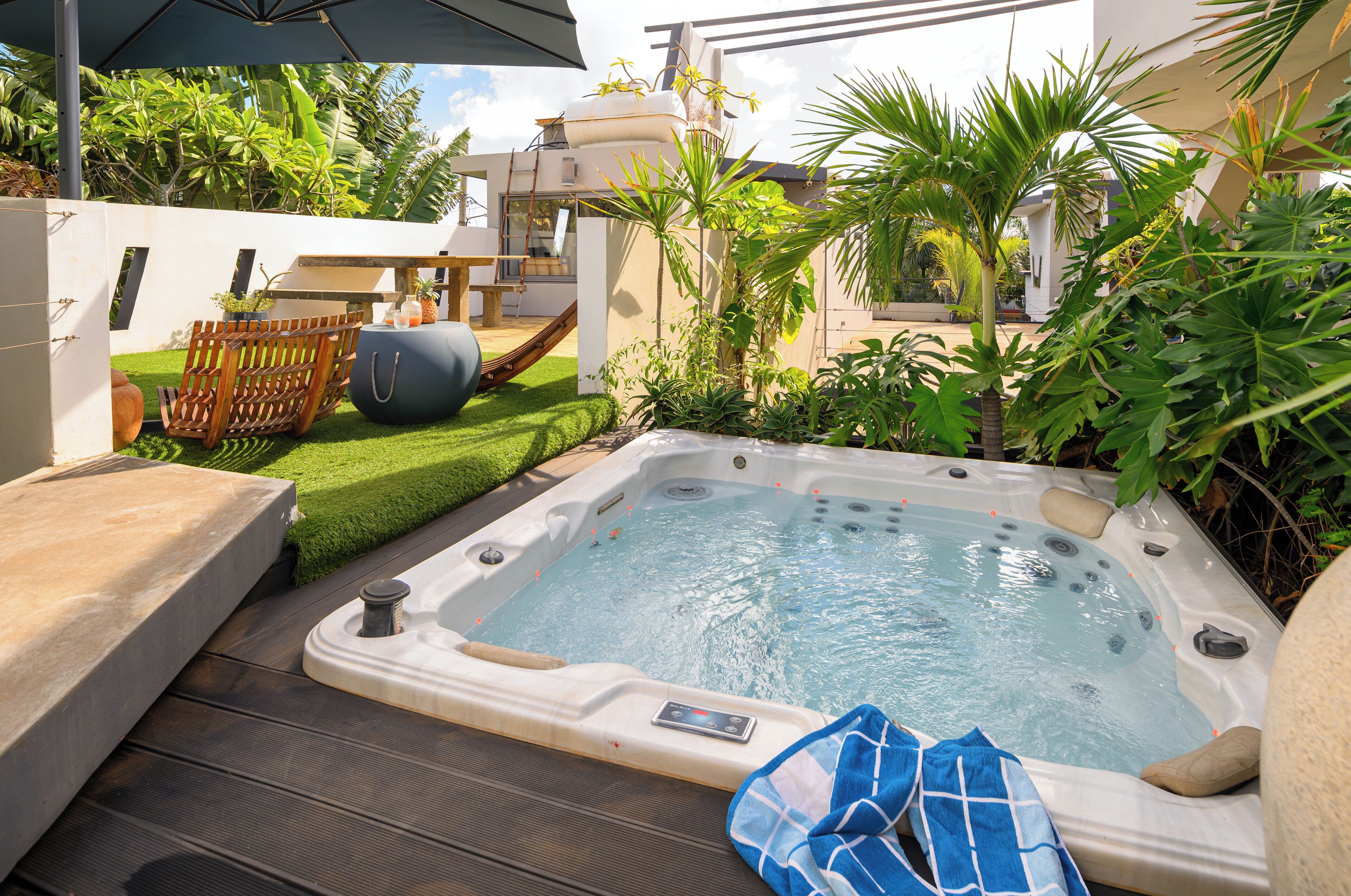Un jacuzzi en un patio trasero con terraza de madera, un tobogán y plantas tropicales.