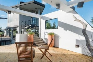 A modern outdoor seating area with a wooden bench and chair, a staircase leading to an upper level, and large potted plants.