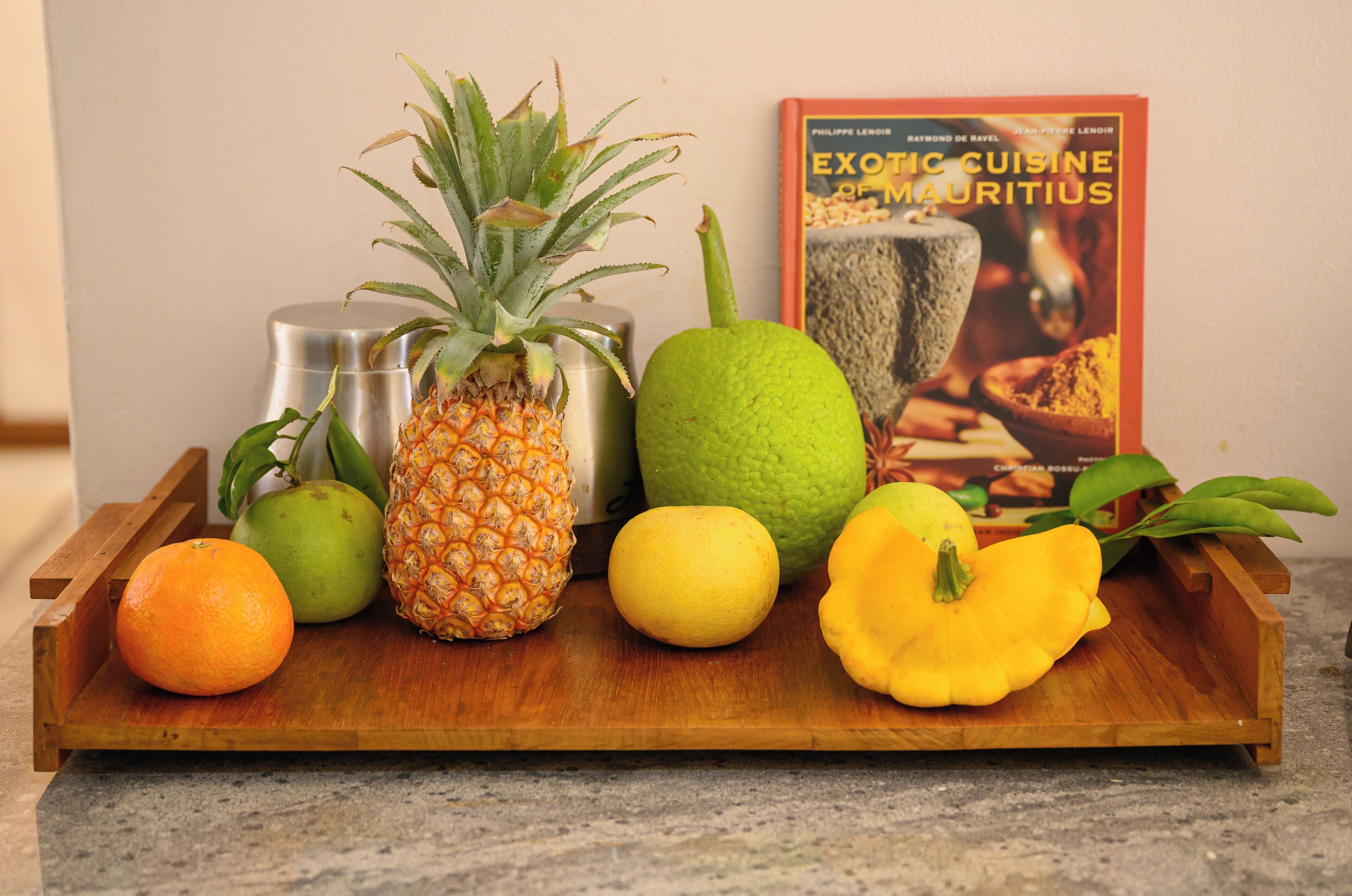 A wooden tray with fruits and a book titled 'Exotic Cuisine of Mauritius' on a kitchen counter.