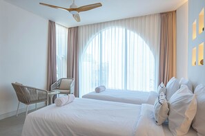 Habitación de hotel con dos camas, una silla, ventilador de techo y ventanas grandes con cortinas.