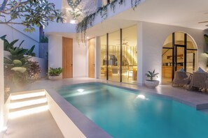 Un área de piscina interior moderna con una piscina cristalina, una puerta de madera y una zona de estar con sillas.