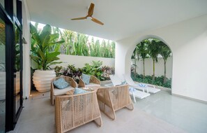 Un espace de détente moderne, doté de meubles en osier, d’un ventilateur de plafond et offrant une vue sur une végétation tropicale.