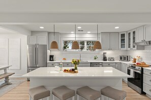 Una cocina moderna con armarios blancos, una isla grande, luces colgantes y electrodomésticos de acero inoxidable.