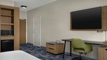 Ein Hotelzimmer mit einem Bett, einem Schreibtisch mit Stuhl, einer Mikrowelle, einem Kühlschrank, einem Kleiderschrank und einem großen Flachbildfernseher an der Wand.