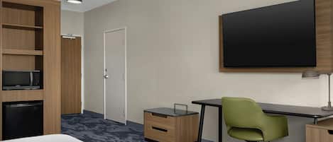Ein Hotelzimmer mit einem Bett, einem Schreibtisch mit Stuhl, einer Mikrowelle, einem Kühlschrank, einem Kleiderschrank und einem großen Flachbildfernseher an der Wand.