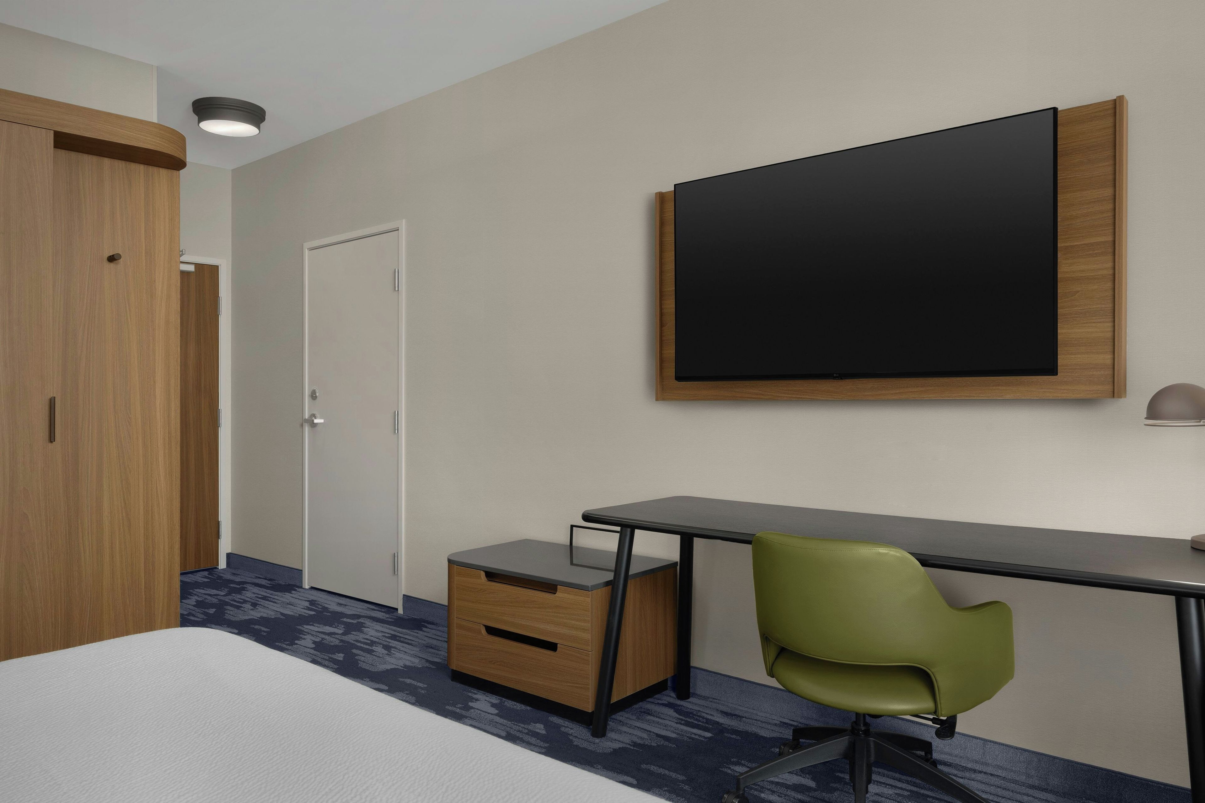 Een hotelkamer met een bed, een bureau met stoel, een kledingkast en een grote flatscreen televisie aan de muur.