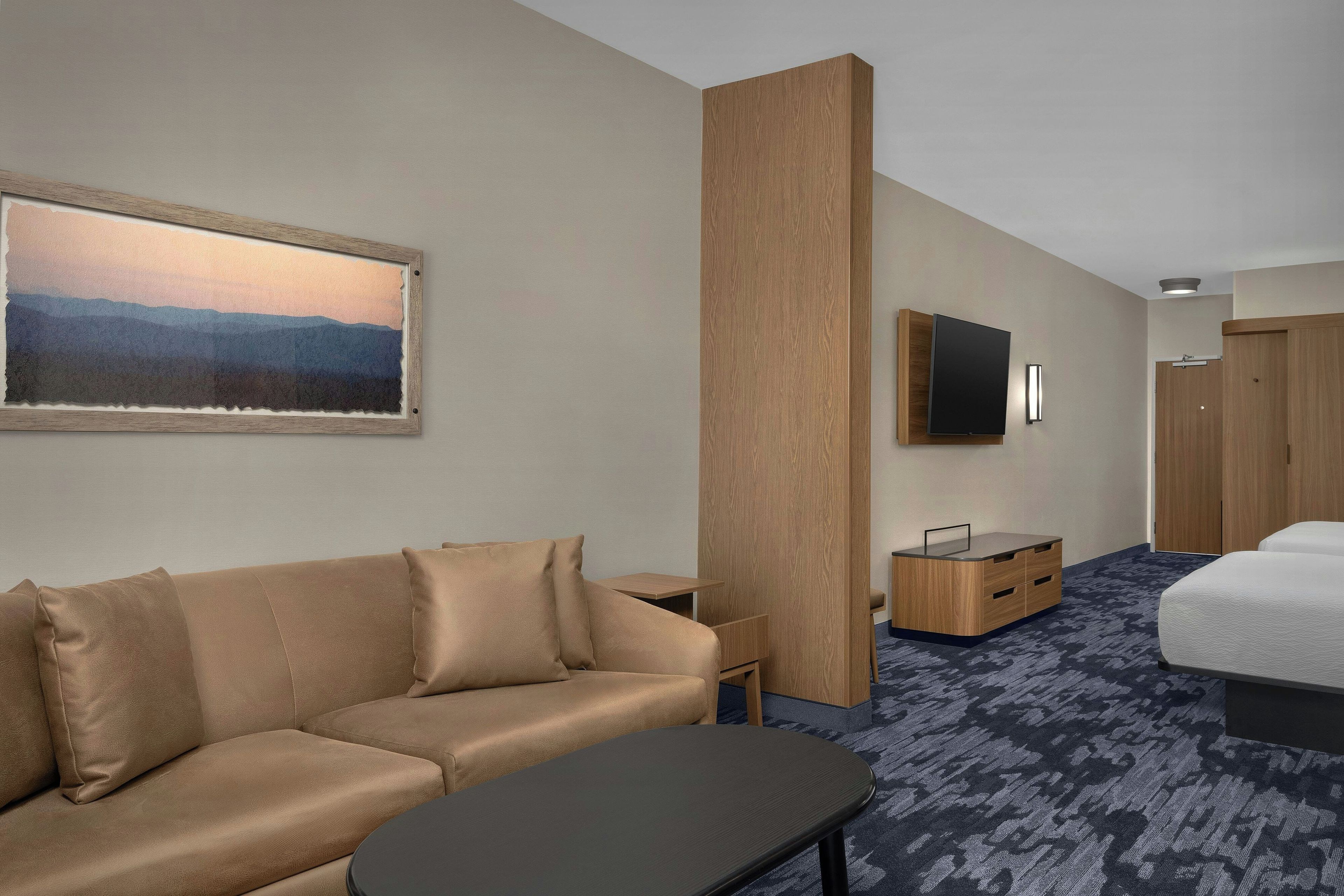 A hotel room with a sofa, a small table, a TV mounted on the wall, a bed, and a wooden wardrobe.