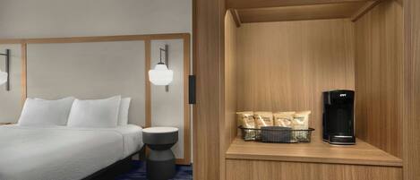 A hotel room with a bed, bedside table, lamp, and a coffee machine on a wooden shelf.