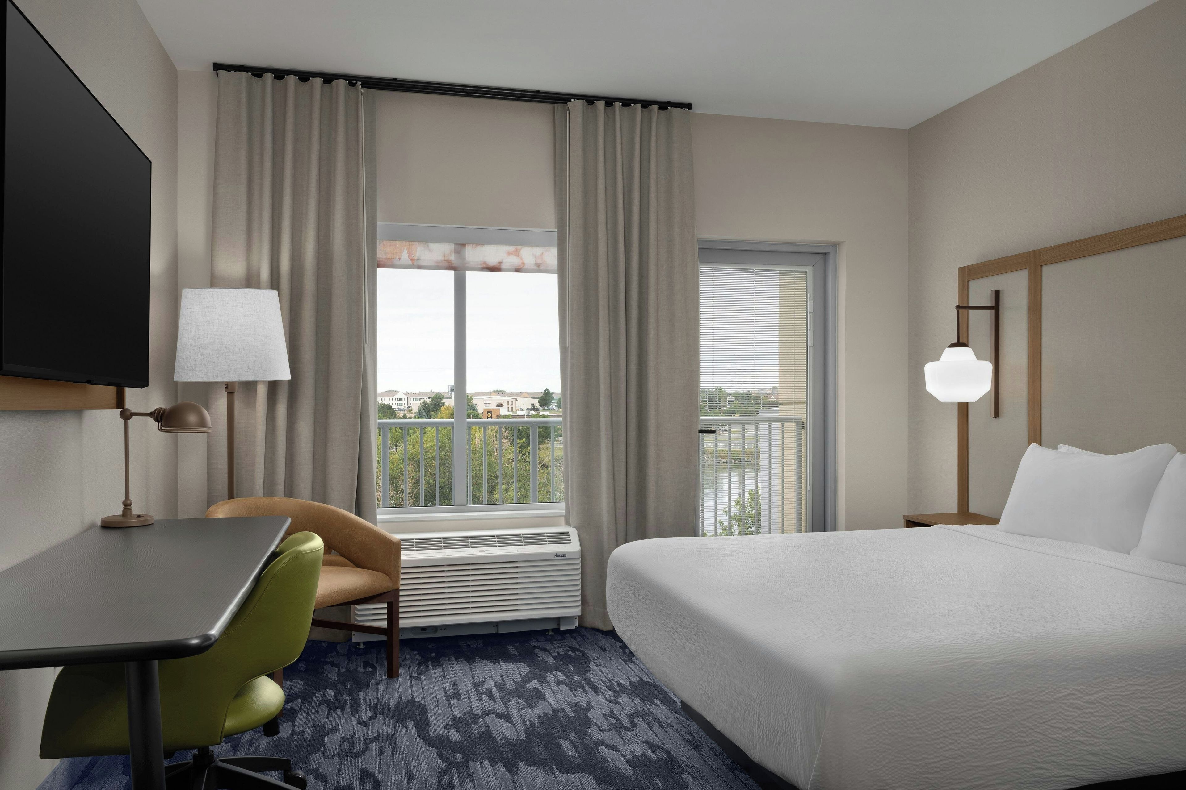 Habitación de hotel con cama, escritorio, silla, televisor y ventana con cortinas, que ofrece vistas al exterior.