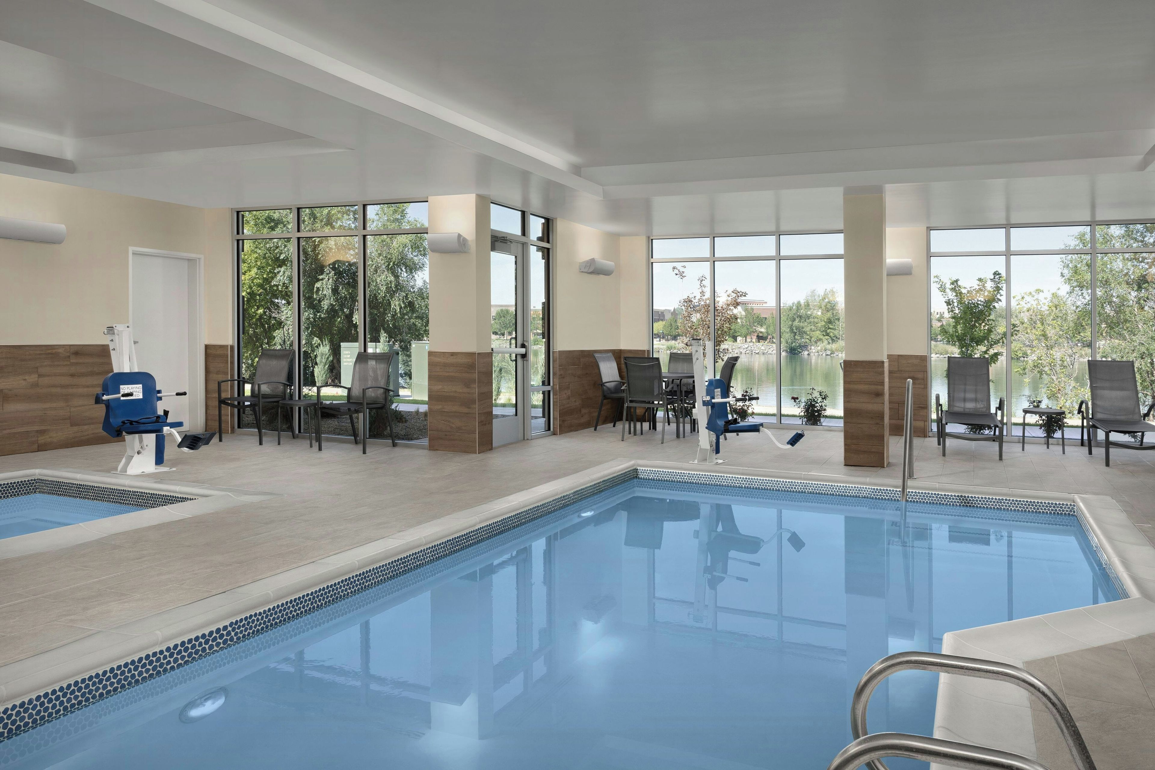 An indoor swimming pool with clear glass walls, lounge chairs, and a view of the outdoors.