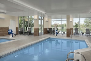 En indendørs swimmingpool med store glasvægge, liggestole og udsigt over naturen.