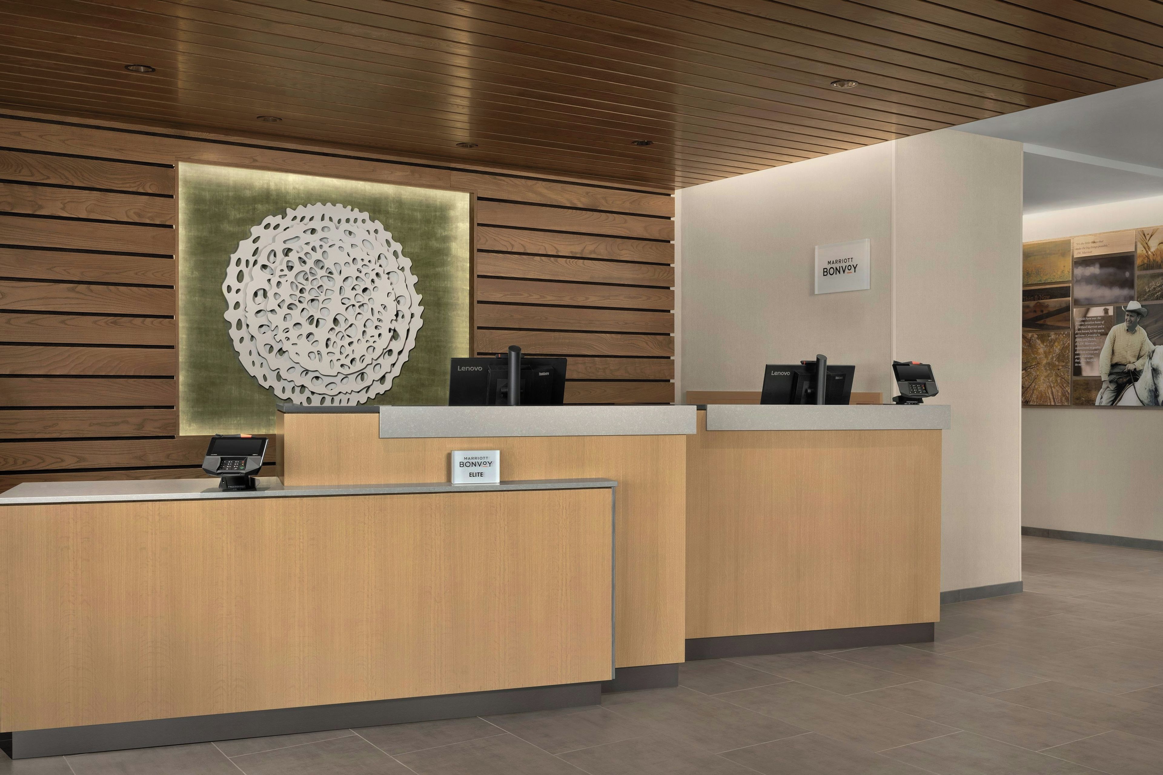A hotel reception desk with a wooden finish, a large decorative wall piece, and a sign indicating the hotel name.