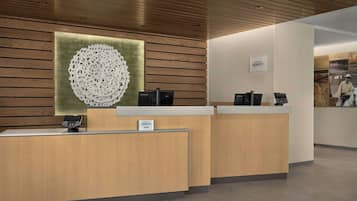 A hotel reception desk with a wooden finish, a large decorative wall piece, and a sign indicating the hotel name.