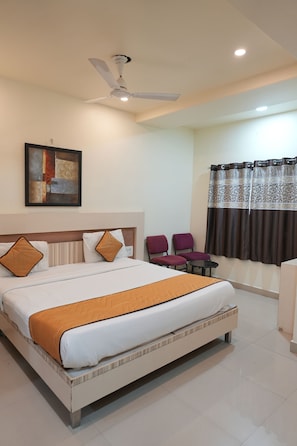 A hotel room with a bed, a ceiling fan, a painting, a window with curtains, and two chairs.
