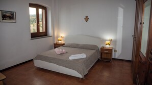 Un dormitorio con cama, dos mesitas de noche con lámparas, una ventana, una puerta de madera y una cruz en la pared.