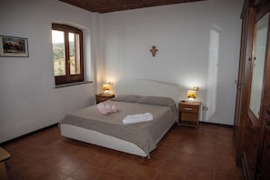 A bedroom with a bed, two bedside tables with lamps, a window, a wooden door, and a cross on the wall. - Cozy Gallelli Castle Farmhouse in Badolato (Calabria)