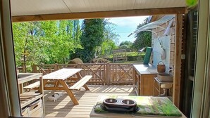 Une vue à travers une fenêtre sur une terrasse en bois, avec des tables de pique-nique, un bol pour chien et un petit jardin.