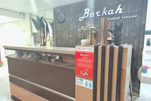 A reception desk with a sign for Barkah Guest House, a clock, and a wooden wall with plants.