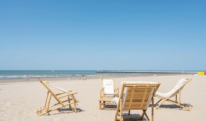 Ein Strand mit zwei Holzliegen, die zum Meer blicken, einem Pier in der Ferne und einem klaren Himmel.