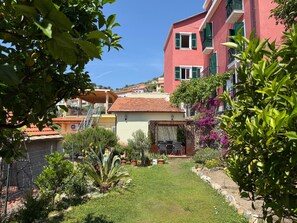 A courtyard with a lawn, potted plants, and a building with a red roof and green shutters. - Apartment 'Rosmarino' with Sea View, Wi-Fi, and Air Conditioning (Moglio)