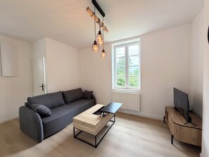 En moderne stue med en grå sofa, et træbord, et fjernsyn på et træstel og et vindue med udsigt til grøn vegetation.