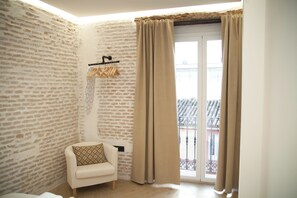 Een kamer met een bakstenen muur, een witte bank met een gekleurd kussen, een kledingrek en een glazen schuifdeur met gordijnen.