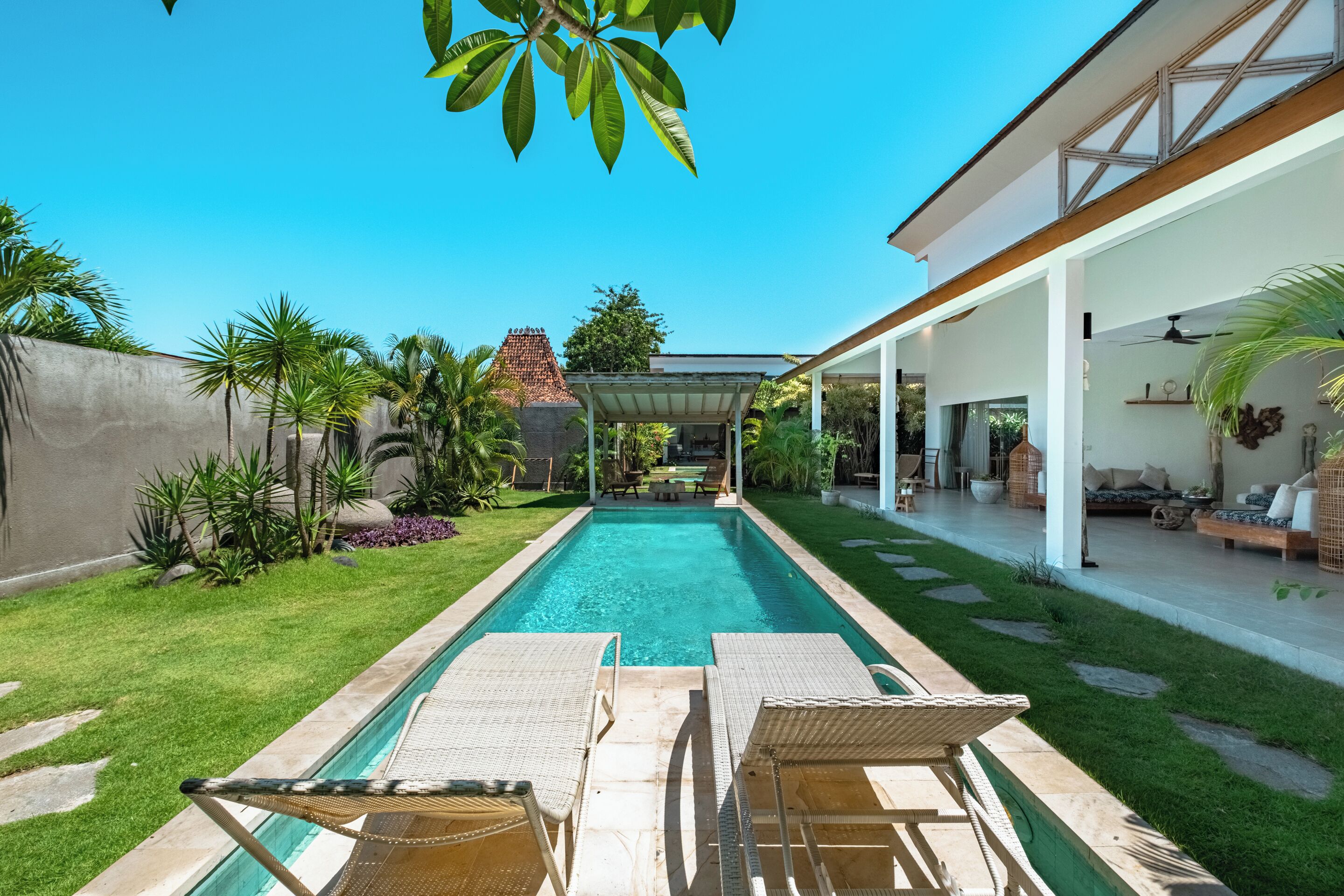 Una piscina con sillas de descanso, un porche techado y un jardín bien cuidado con palmeras.