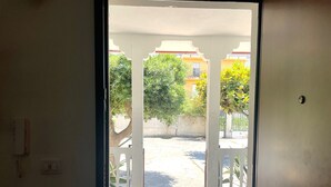 Une porte ouverte donne sur un espace extérieur dallé, avec un portail blanc, un arbre et un bâtiment en arrière-plan.