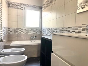 Een moderne badkamer met een badkuip, twee hygiënespotten, een raam en een betegelde spatwand.