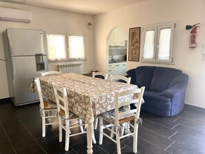 A dining area with a table set for four, a refrigerator, a water dispenser, a blue armchair, and a kitchen in the background.