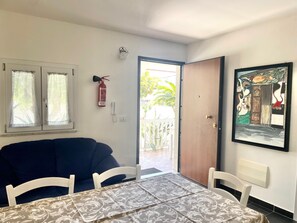 A room with a sofa, a table with a patterned tablecloth, a fire extinguisher, a phone, and a door leading outside.