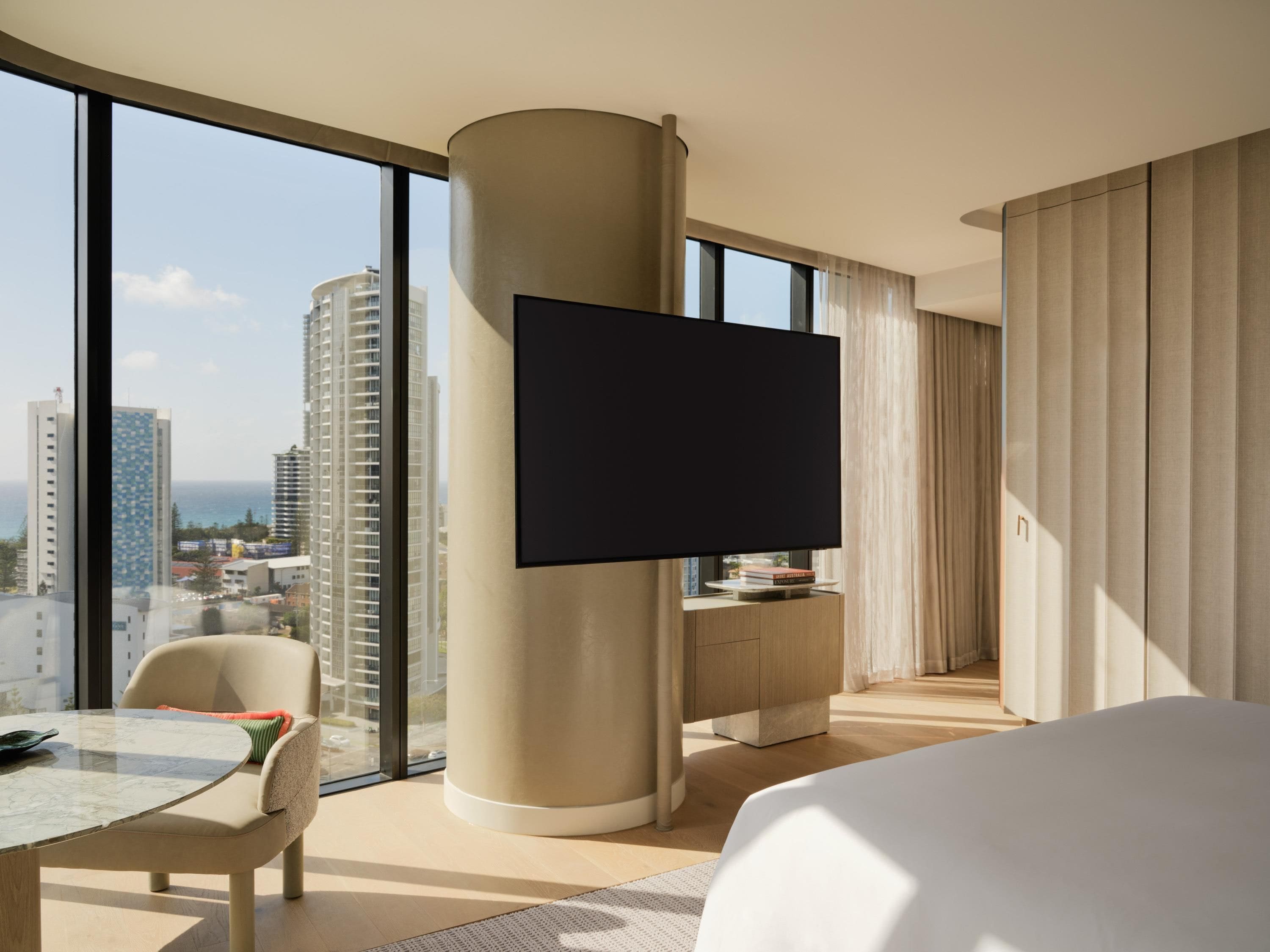 A hotel room with a large window, a flat-screen TV, a bed, a dining table, and a view of the cityscape.