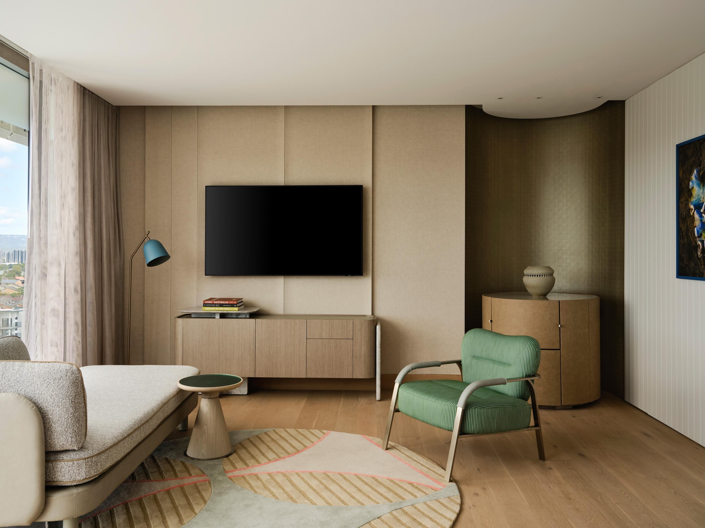 A modern hotel room with a flat-screen TV, a green upholstered chair, a small round table, and a bookshelf with books.