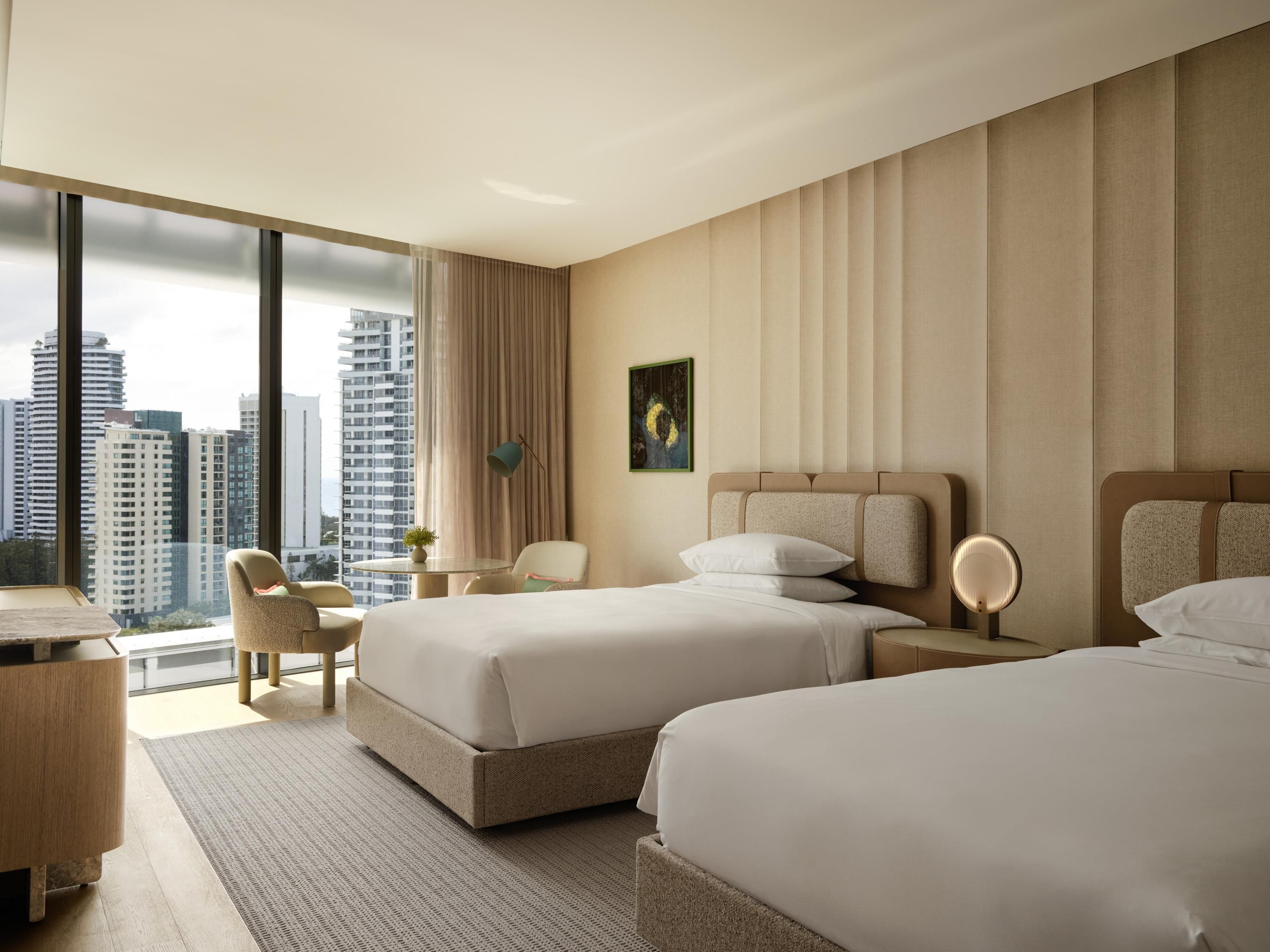 A hotel room with two beds, a desk, a chair, a small table, and a view of tall buildings.