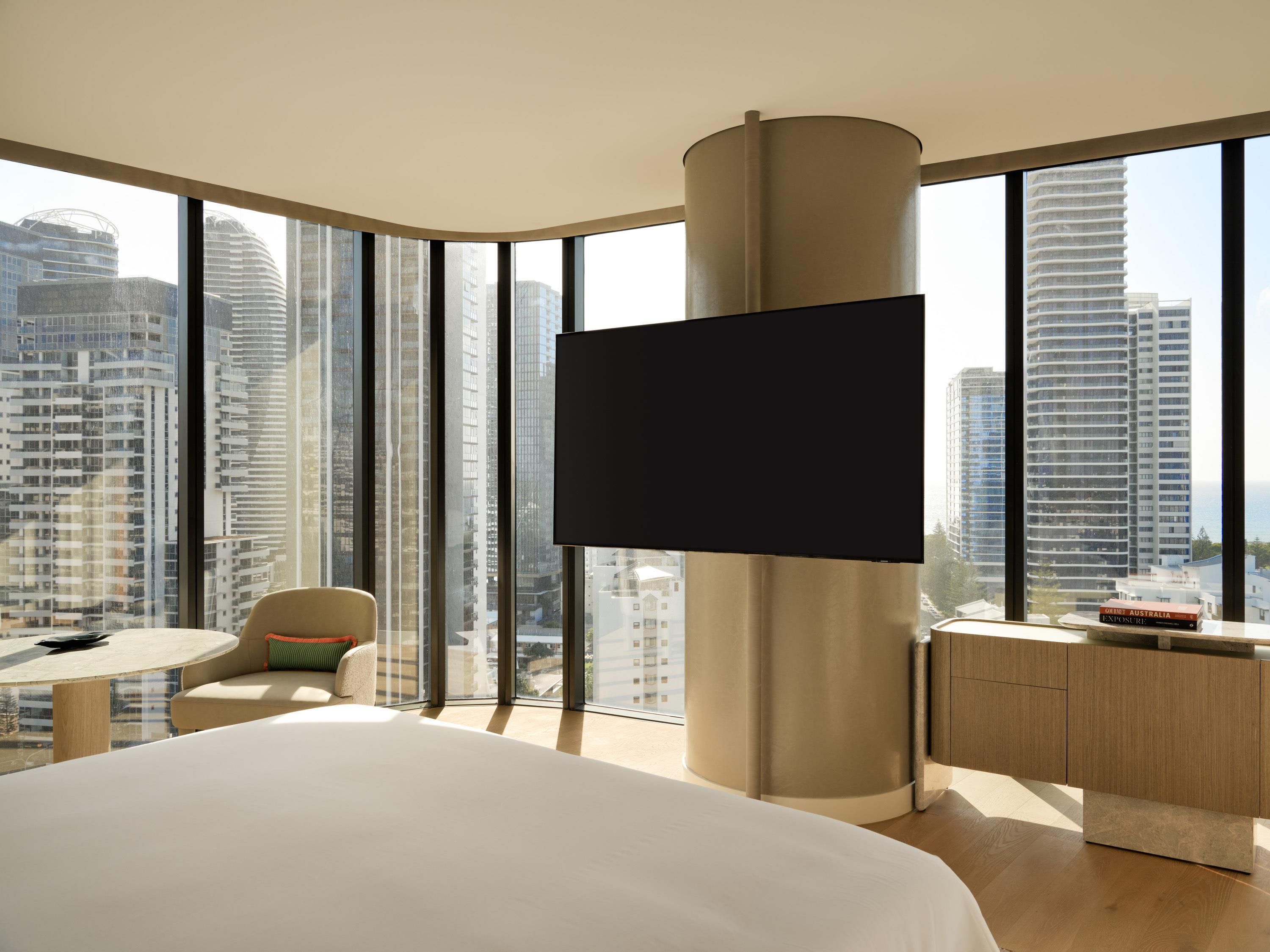 A modern hotel room with a large bed, a flat-screen TV mounted on a column, a small round table with a chair, and a view of tall buildings through large windows.