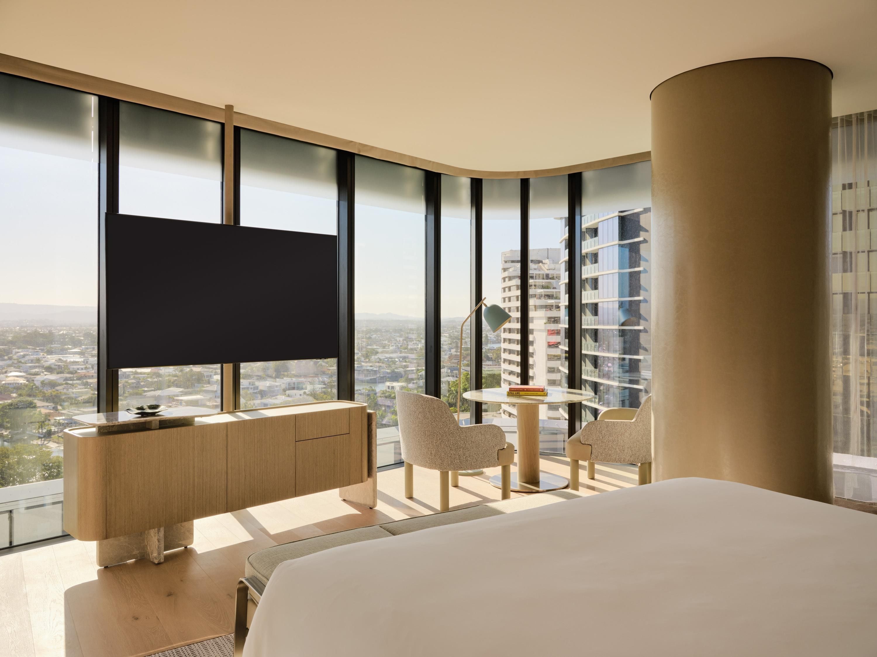A modern hotel room with a large bed, a desk, two chairs, a small table, and a large window offering a city view.