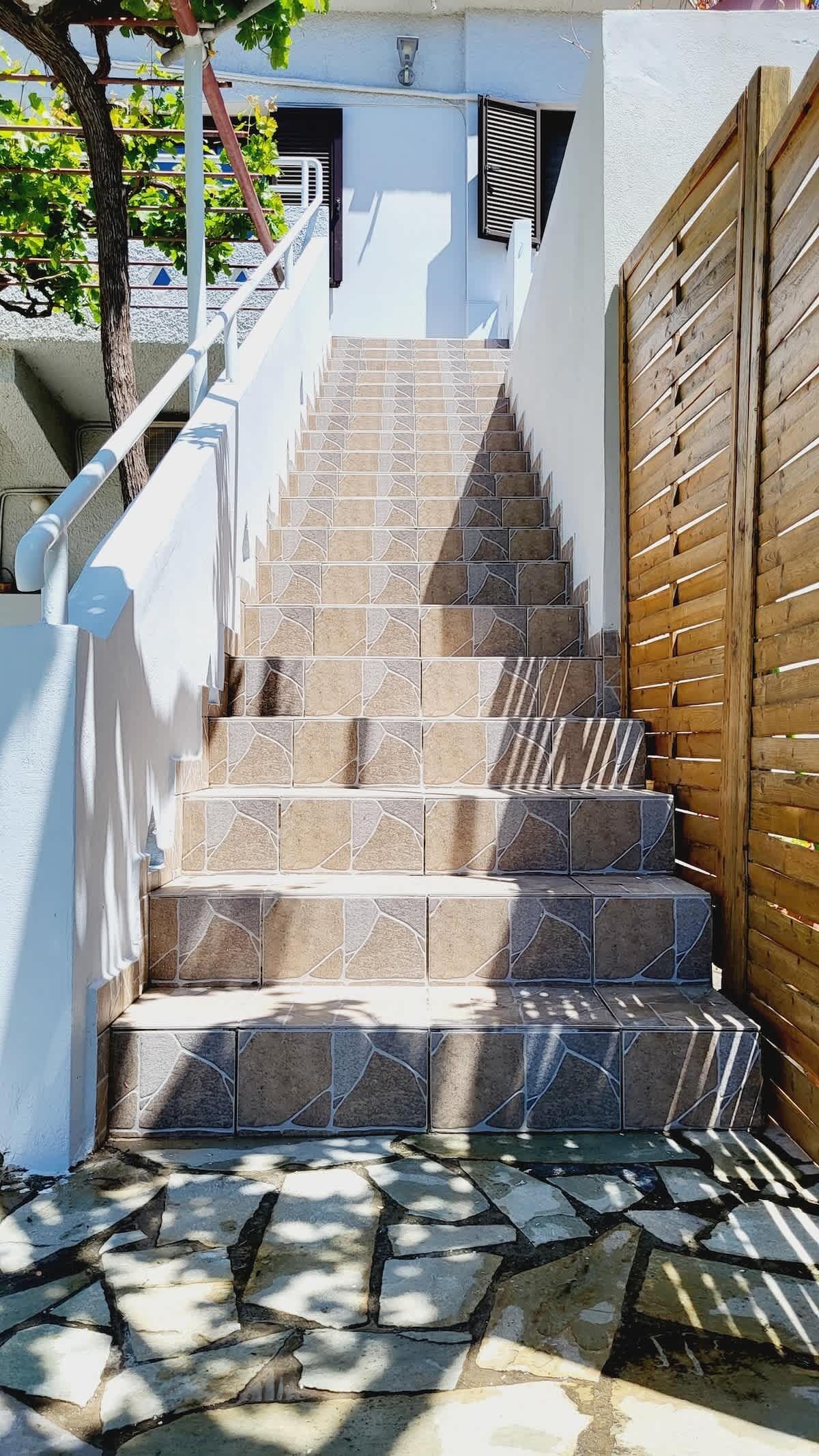 A set of stone-tiled stairs leading to a wooden door, with a white wall and a metal railing on the left.